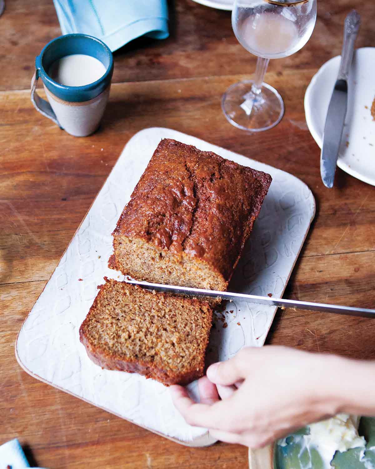 A person cutting a slice from a loaf of whole wheat banana bread on a white cutting board.