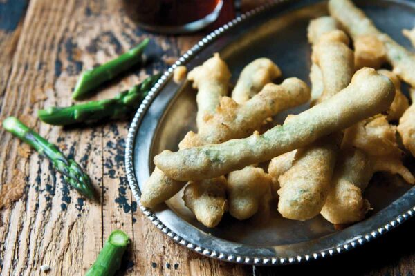 A silver platter filled with asparagus tempura with some pieces of asparagus and a glass of beer on a wooden table.