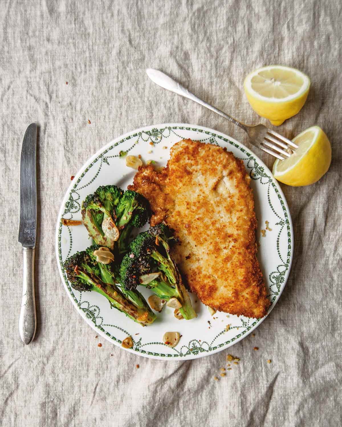 A piece of bread crumb coated chicken on a patterned plate with broccoli and a fork and two lemon halves on the side.