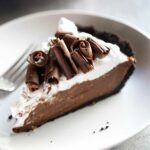 A slice of chocolate cream pie, topped with chocolate curls on a white plate with a fork beside it.
