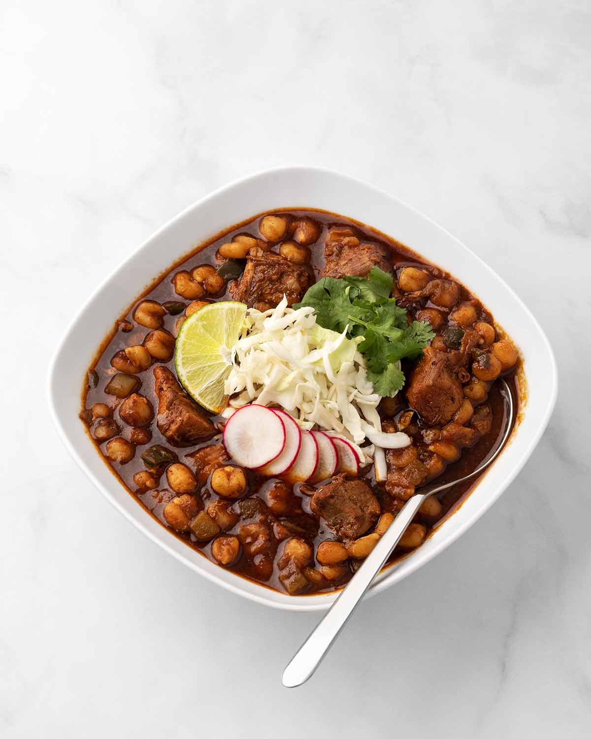 A square white bowl filled with Instant Pot pork pozole, garnished with radish, shredded cheese, lime, and cilantro, with a spoon resting in it.