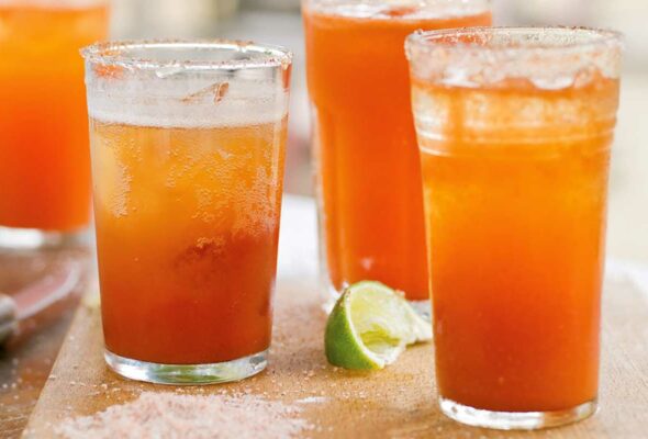 Four glasses of michelada on a wooden board with a lime wedge in between them.