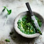 A stone bowl of mint pesto with a knife on top on a metal sheet pan