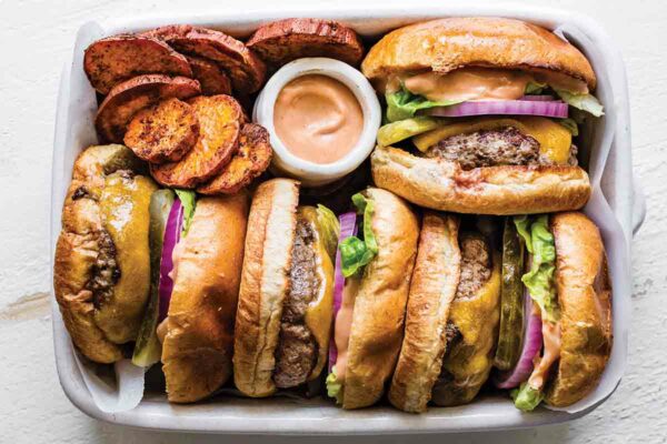Four cheeseburgers with everything sauce tucked into a white casserole dish with sweet potato chips and a dish of everything sauce.