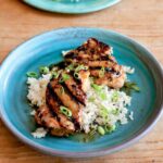 Two pieces of grilled lemongrass chicken on top of rice with scallion garnish on a blue plate.