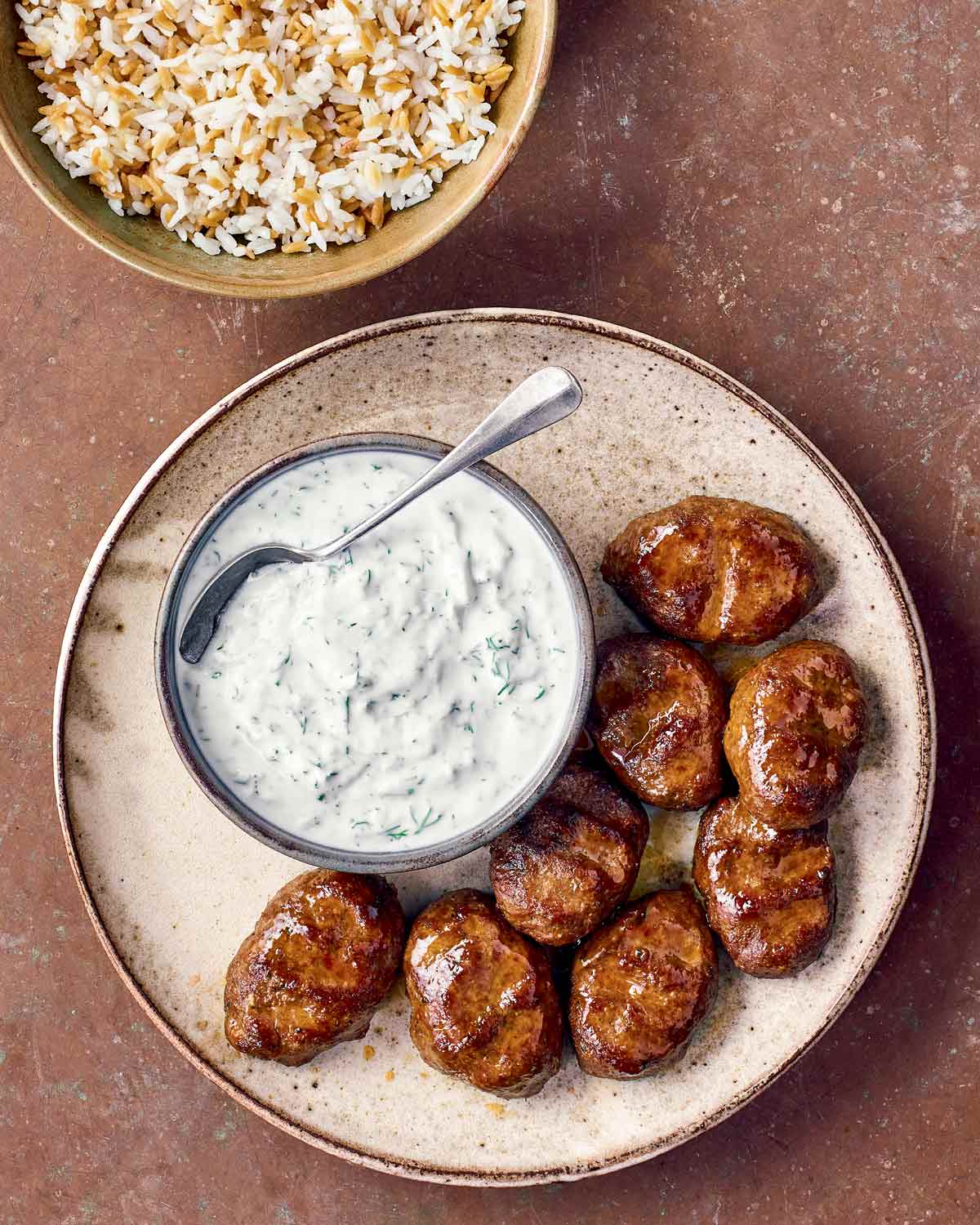 Eight kafta, or spiced kebabs, on a plate with a bowl of preserved lemon yogurt next to it and a bowl of rice beside it.