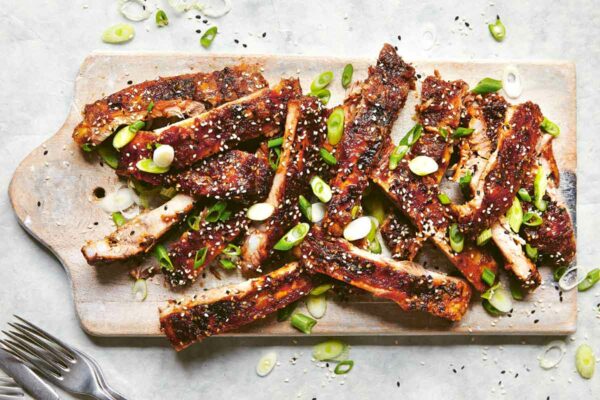 A pile of Korean-style BBQ ribs on a wooden board, garnished with scallions and sesame seeds with several forks on the side.