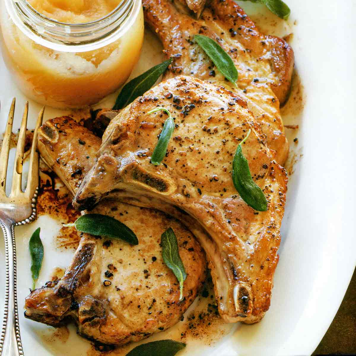 A white platter with four pork chops with applesauce in a jar on the side and fried sage leaves sprinkled over the pork chops.