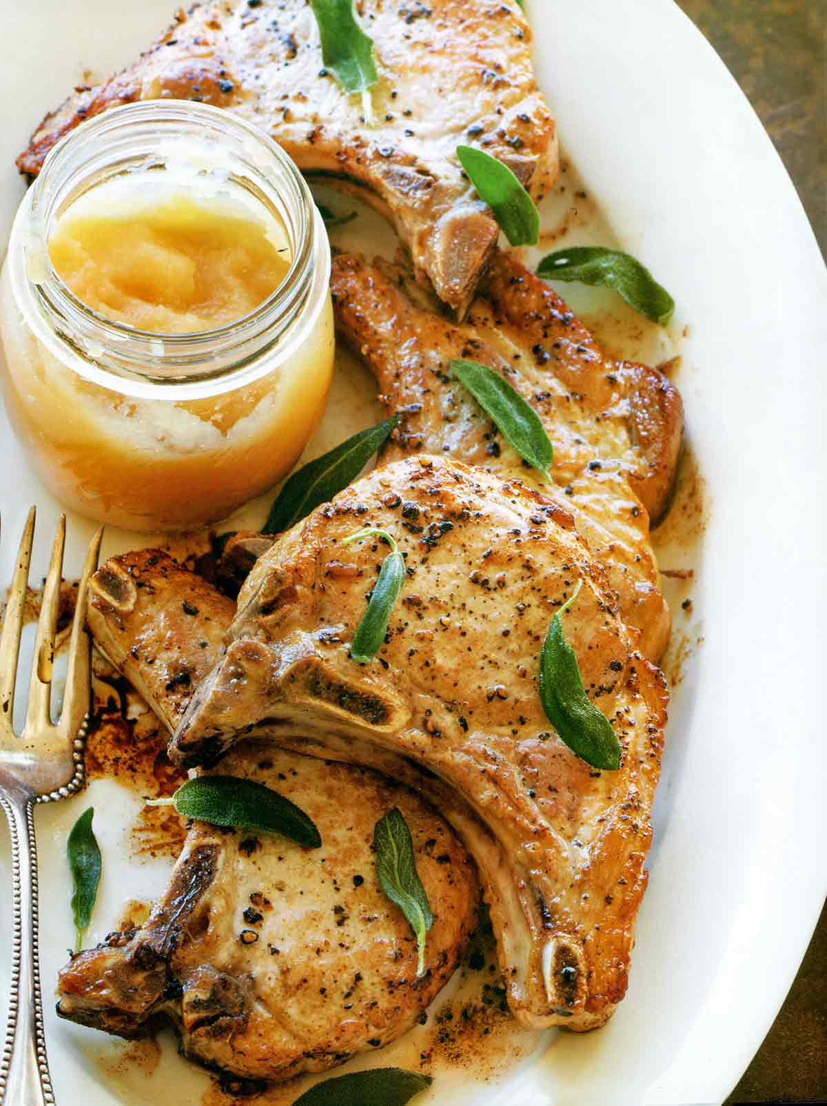 A white platter with four pork chops with applesauce in a jar on the side and fried sage leaves sprinkled over the pork chops.