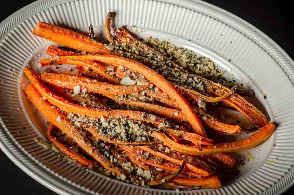 A white oval platter topped with roasted carrots with dukkah and nuts sprinkled on top.