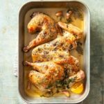 A spatchcock roast chicken in a deep roasting pan with sprigs of thyme, lemon halves, and garlic cloves.
