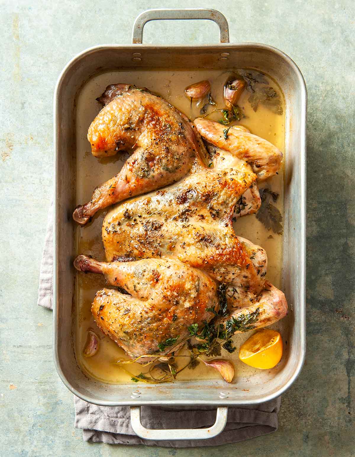 A spatchcock roast chicken in a deep roasting pan with sprigs of thyme, lemon halves, and garlic cloves.