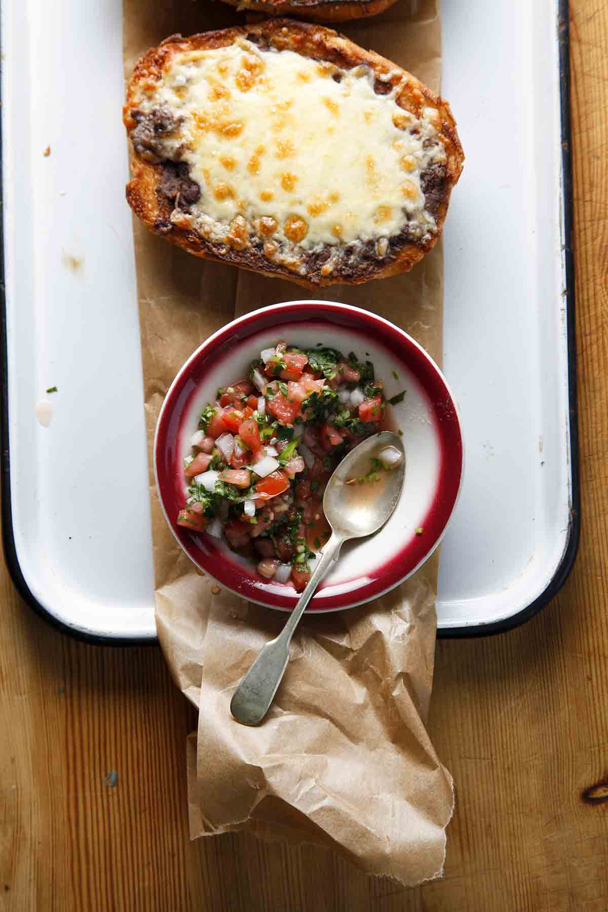 Two people reaching for traditional molletes on a platter with a bowl of salsa in the middle.