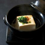 A black bowl with a block of warm tofu with soy sauce, ginger, and scallion garnish.