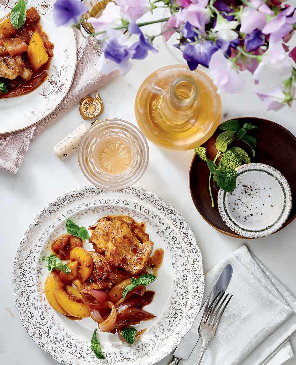 A patterned plate topped with chicken thighs with spicy peach sauce on a table with a bottle and glass of wine, flowers, cutlery, and a napkin.