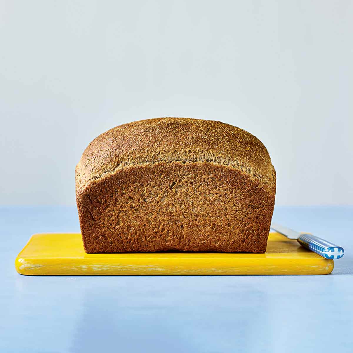 A loaf of unsliced whole wheat bread sitting on a yellow cutting board with a blue-handled knife beside it