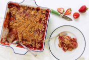 A square baking dish filled with strawberry rhubarb crumble with balsamic drizzle, with a portion on a plate and a few strawberries on the side.