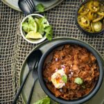 A bowl of Texas-style chili with pork and brisket with a fork, spoon, and bowls of lime, cilantro, and pickled jalapenos on the side.