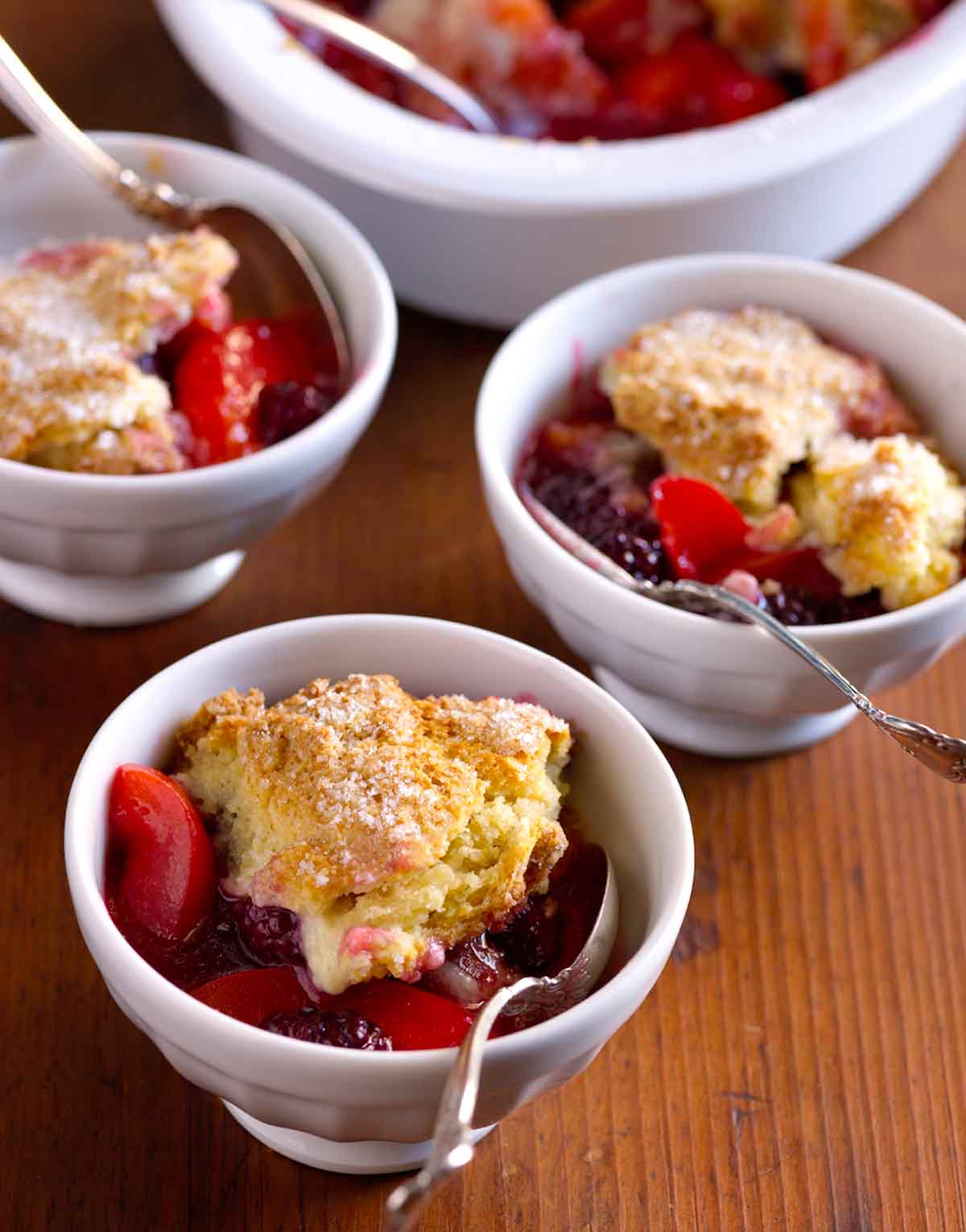 Small white bowls with spoons, filled with apricot and blackberry compote, coered with dollops of sugared biscuit topping.