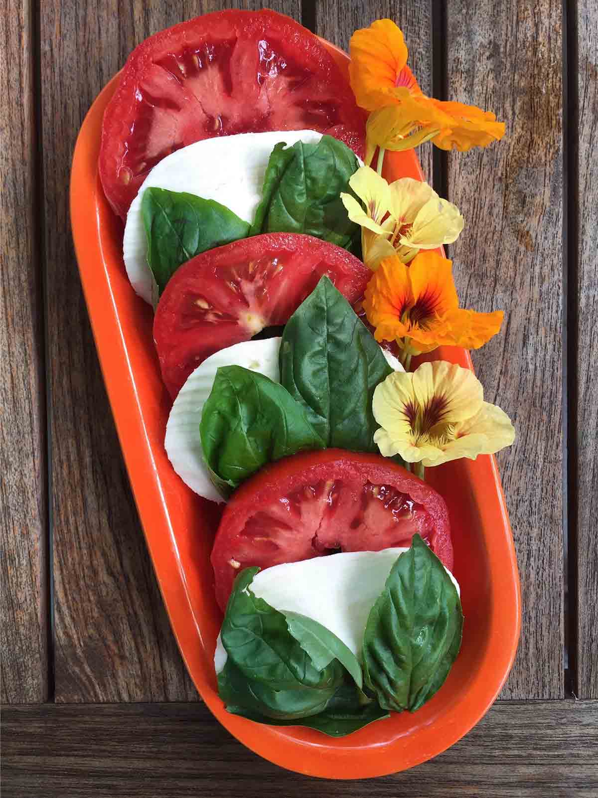 A caprese salad of tomato, mozzarella, and basil, garnished with flowers on a red platter.
