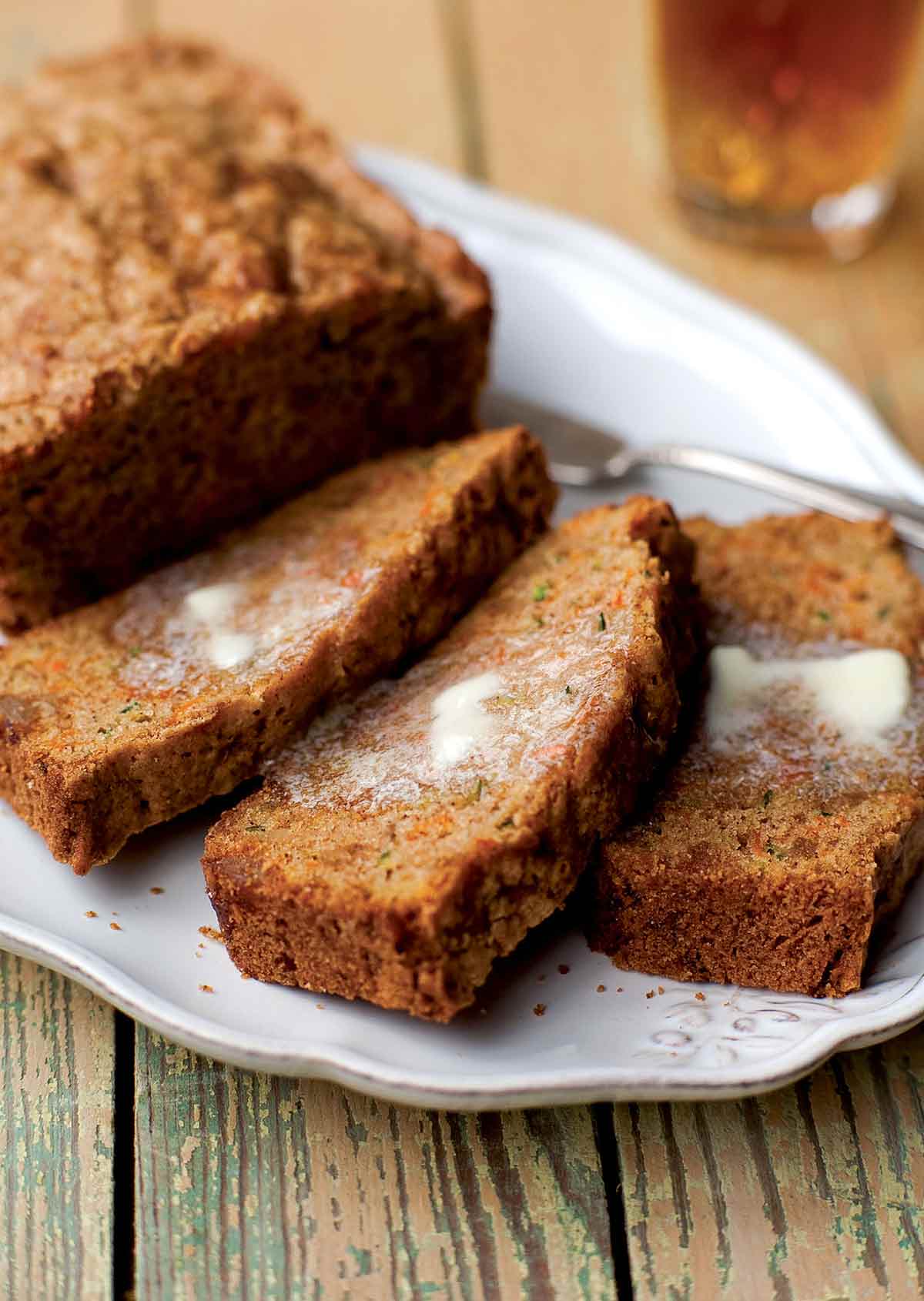 A loaf of carrot zucchini bread with candied ginger on a white platter with three slices cut from it and buttered.