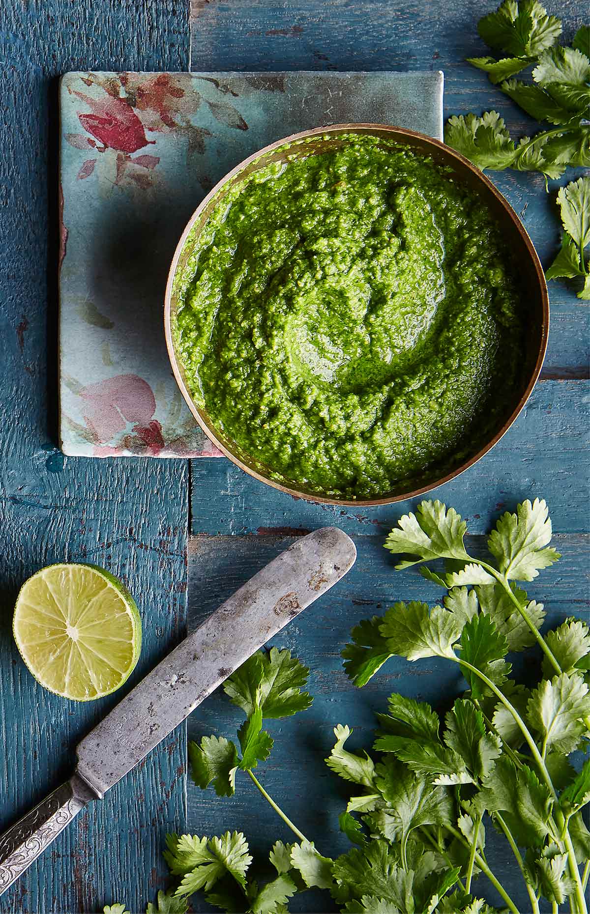 A bowl of bright green cilantro chutney on a blood wooden table with a flower napkin, cilantro leaves, half a lime, and a knife.