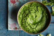 A bowl of bright green cilantro chutney on a blood wooden table with a flower napkin and cilantro leaves.
