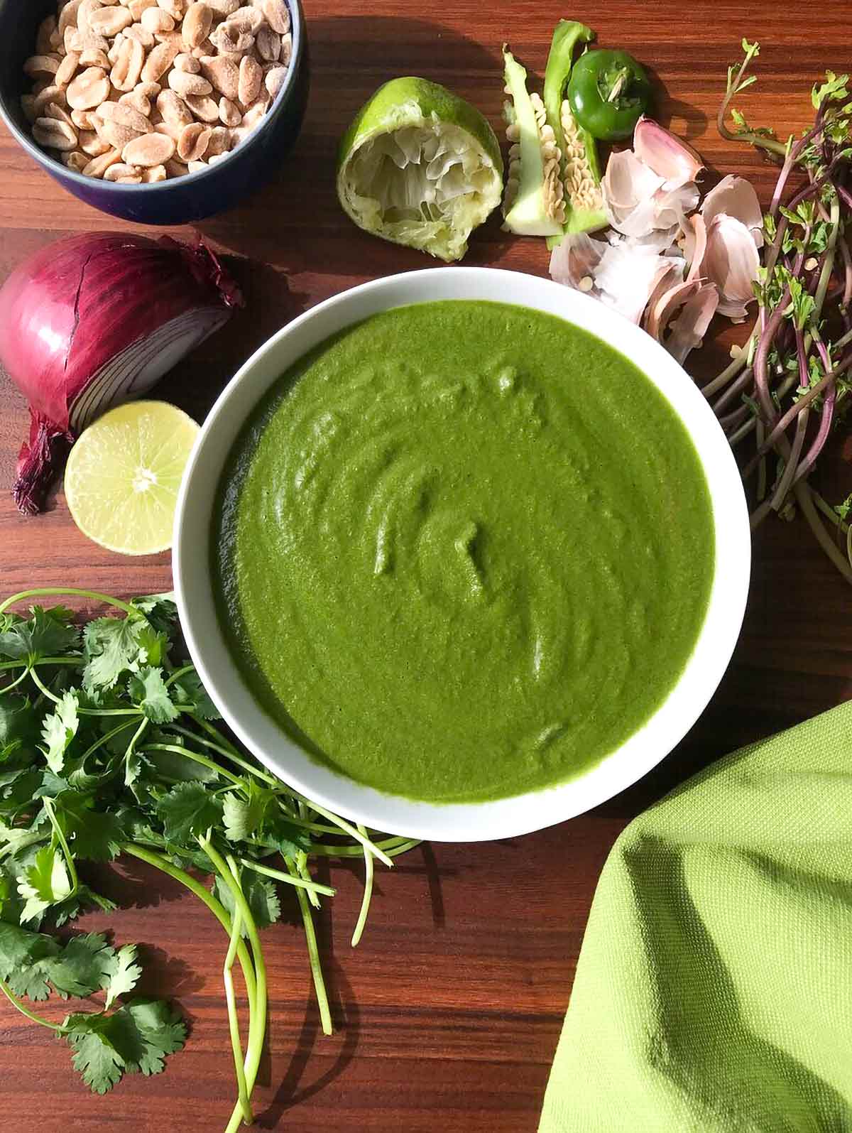 A white bowl of green chutney on a wooden table with a bowl of peanuts, cilantro, limes, garlic, and onions.