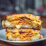 A white bowl on a wooden table, with a grilled cheese sandwich inside filled with cheese, Doritos, bacon, and guacamole.