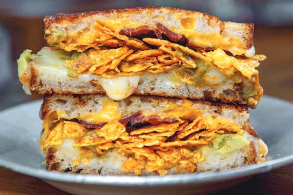 A white bowl on a wooden table, with a grilled cheese sandwich inside filled with cheese, Doritos, bacon, and guacamole.