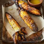 Three cobs of grilled corn with husks attached, lying on a metal sheet pan with parchment paper.