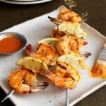 Two skewers of grilled shrimp with piri-piri sauce in a dish on the side.