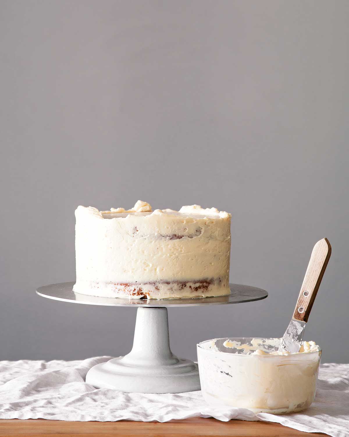 A partially frosted cake on a cake stand with a bowl of frosting and spatula on the side.