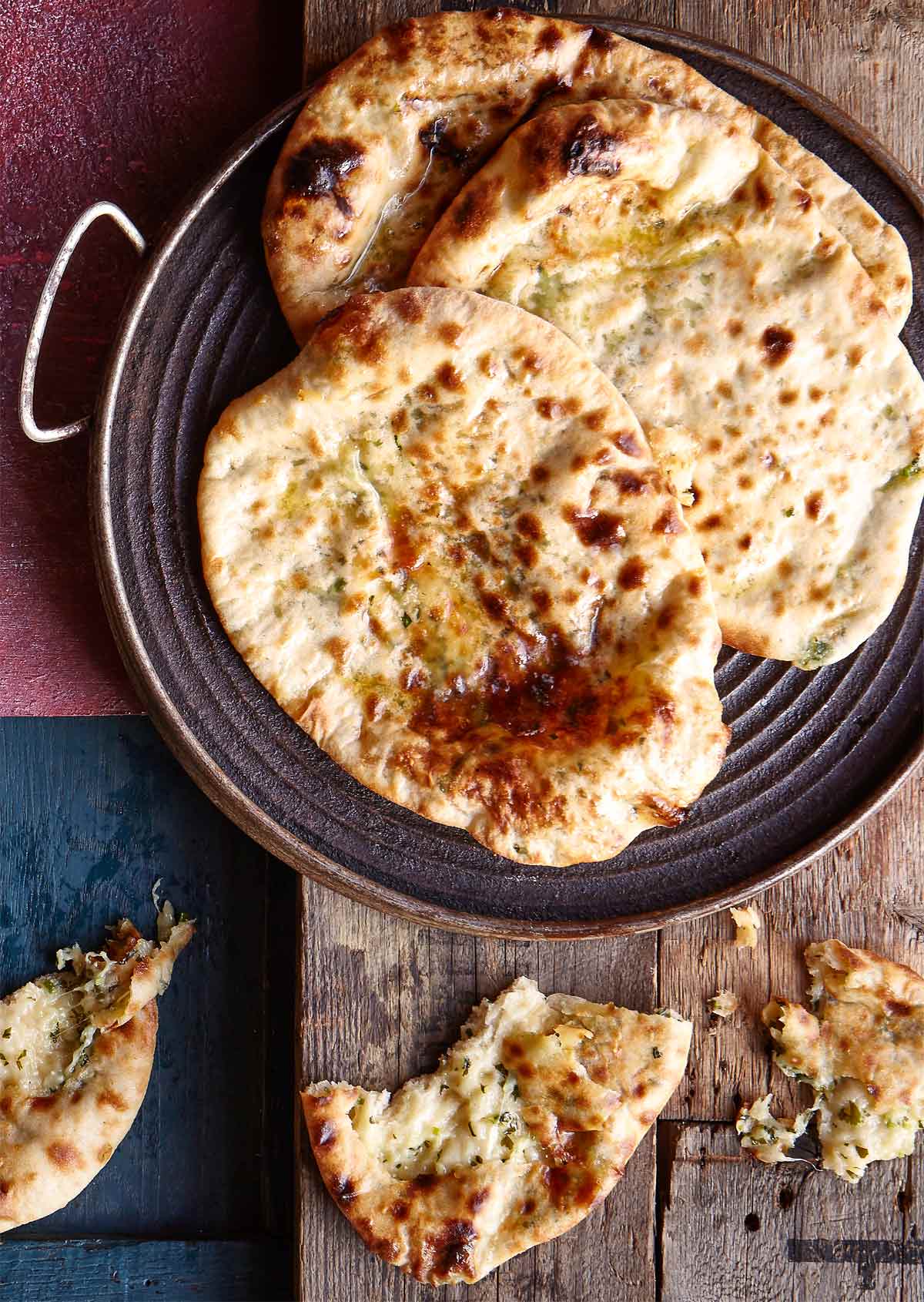 Three cheese and chile naan bread, drizzled with butter, in a metal dish on a wooden table, beside pieces of naan.