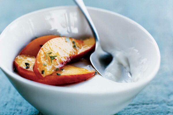 A white bowl filled with panna cotta with peaches, garnished with thyme and a spoon resting inside.