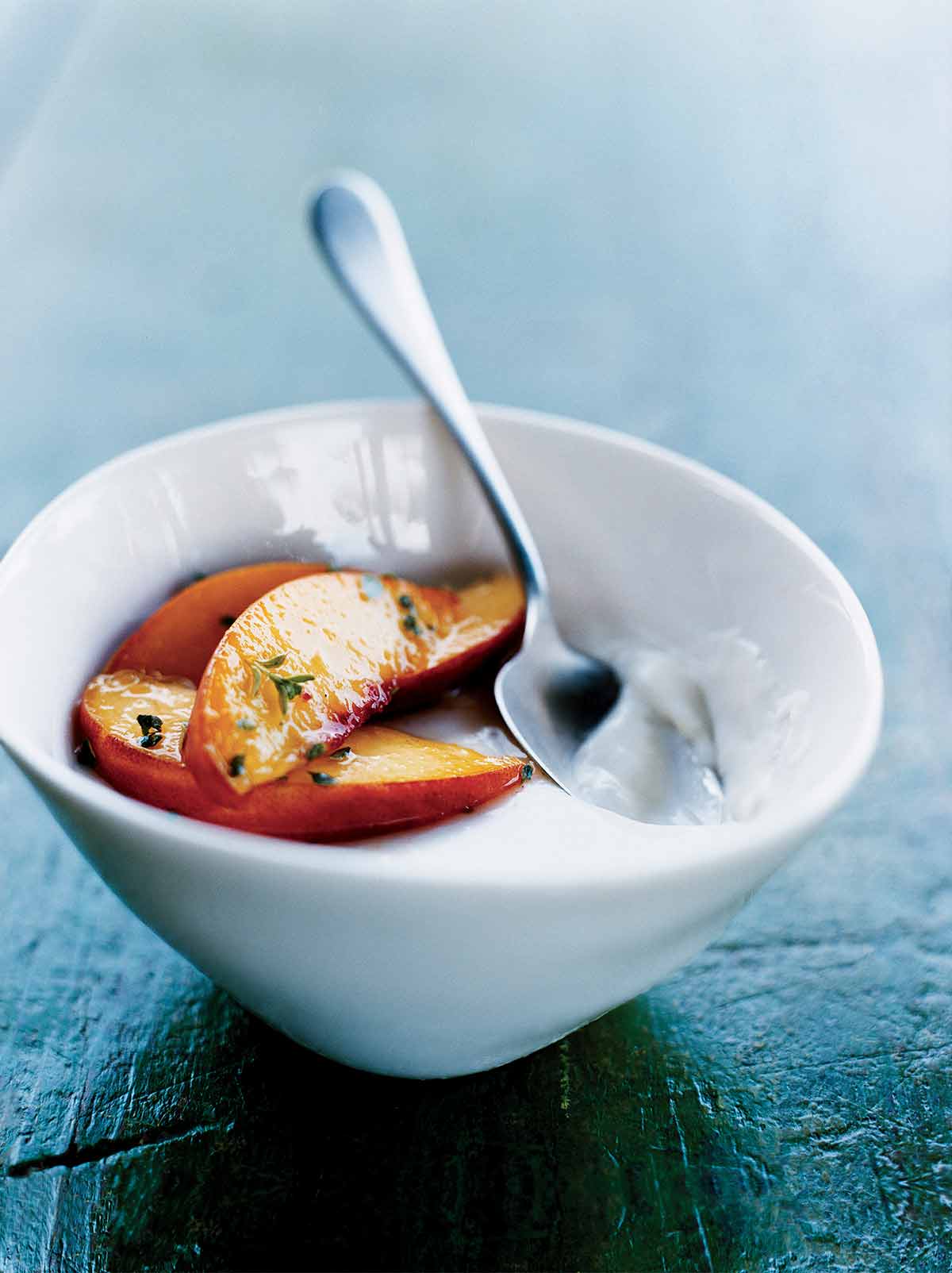 A white bowl filled with panna cotta with peaches, garnished with thyme and a spoon resting inside.