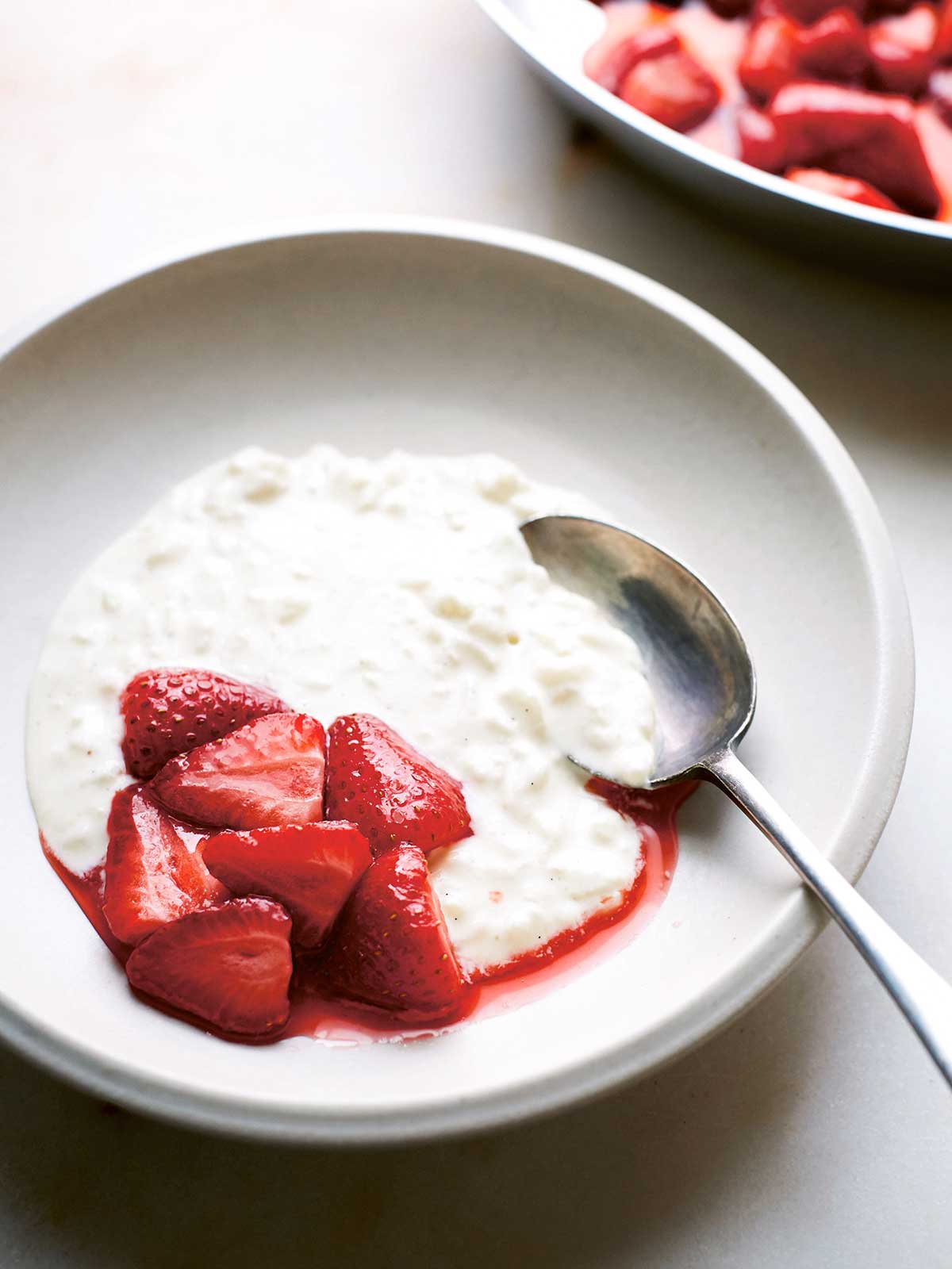 A bowl of rice pudding with a spoon and a scoop of halved strawberries and juice