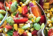 A close up of two serving spoons in a bowl of corn, lima beans, chopped peppers, red onion and cherry tomatoes, dressed with parsley, mint, and dressing.