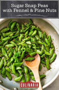 A metal wok filled with halved snap peas, sprinkled with fennel, pine nuts, and lemon zest, with a wooden spoon.