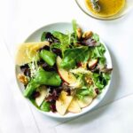 A large white bowl filled with apples, parmesan, walnuts, and mixed green salad, with a small bowl of honey dressing next to it.