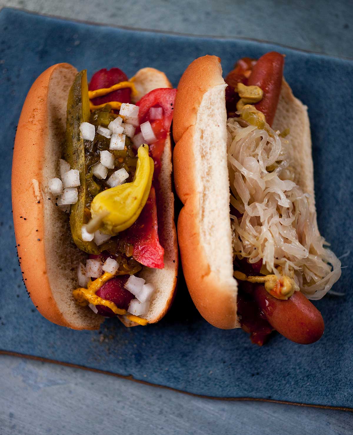 A dark blue pottery plate with 2 hot dogs, one dressed with pickles, onions, tomatoes, mustard, and peppers, the other with mustard and sauerkraut.
