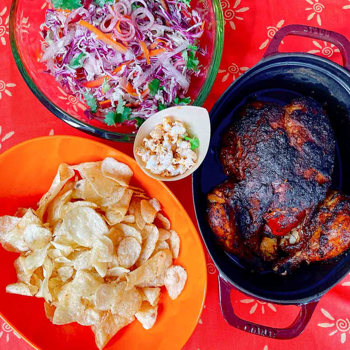 A red tablecloth with an enamel roaster containing a crisp, roasted chicken, an orange bowl of potato chips, a green glass bowl of coleslaw, and a small bowl of popcorn.