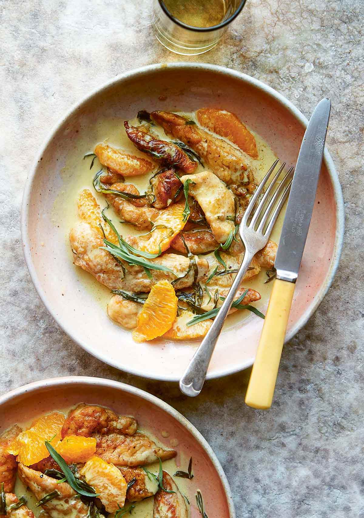 2 bowls of chicken tenders in a cream sauce with slices of orange and chiffonaded tarragon leaves, with a knife and fork.