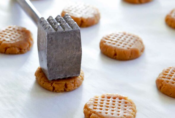 A parchment covered counter with lines of raw peanut butter cookies and a meat-tenderizing mallet.