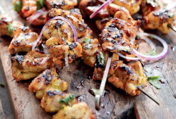 6 grilled chicken skewers piled on a wooden cutting board and sprinkled with slices of red onion and cilantro.