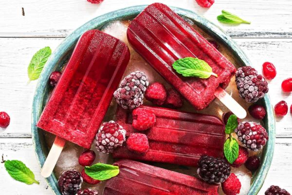 White barn board with a green bowl filled with ice and frozen berries, and 4 deep red raspberry hard seltzer popsicles lying on top, garnished with fresh mint leaves.