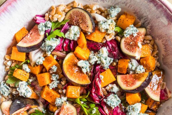 A red and white bowl filled with cubes of squash, shreds of raddichio, slices of figs, crumbles of blue cheese, and farro.