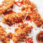 5 panko breaded chicken tenders drizzled with deep red hot sauce and chile slices, with a bowl of more spicy honey and a honey dipper.