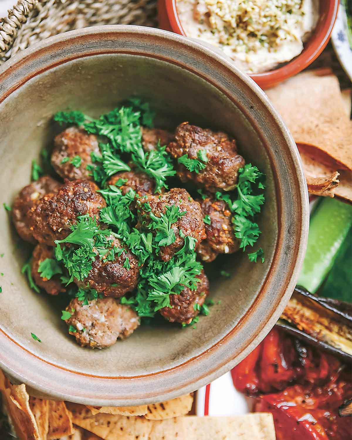 A pottery bowl filled with merguez meatballs, garnished with parsley, surrounded by pitas, hummus, and roasted red peppers.