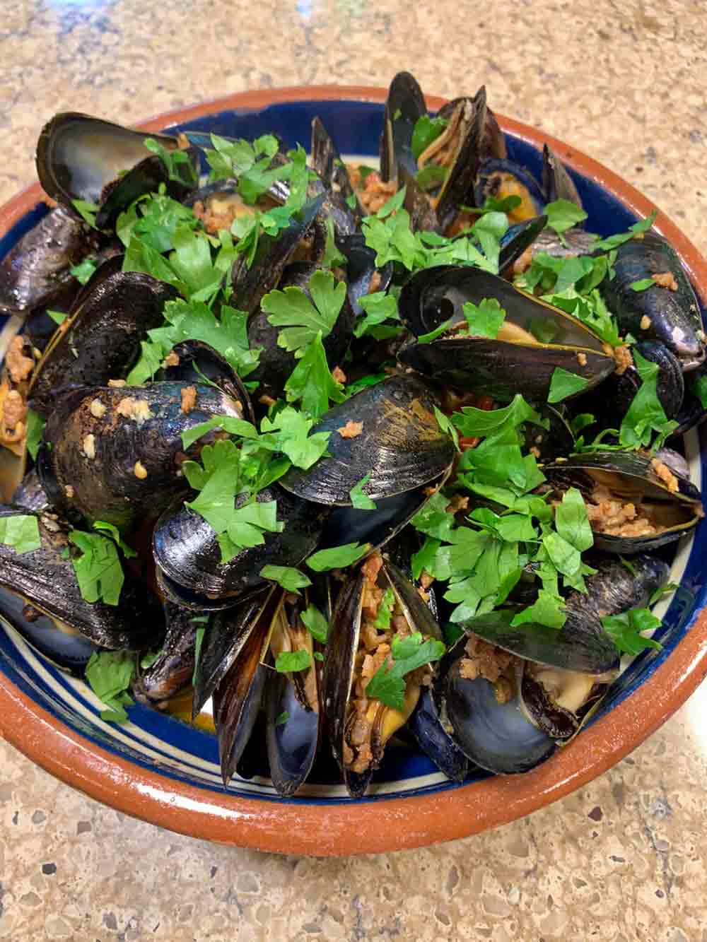 A blue and terracotta bowl full of mussels, tomatoes, chunks of chorizo, all sprinkled with loads of parsley